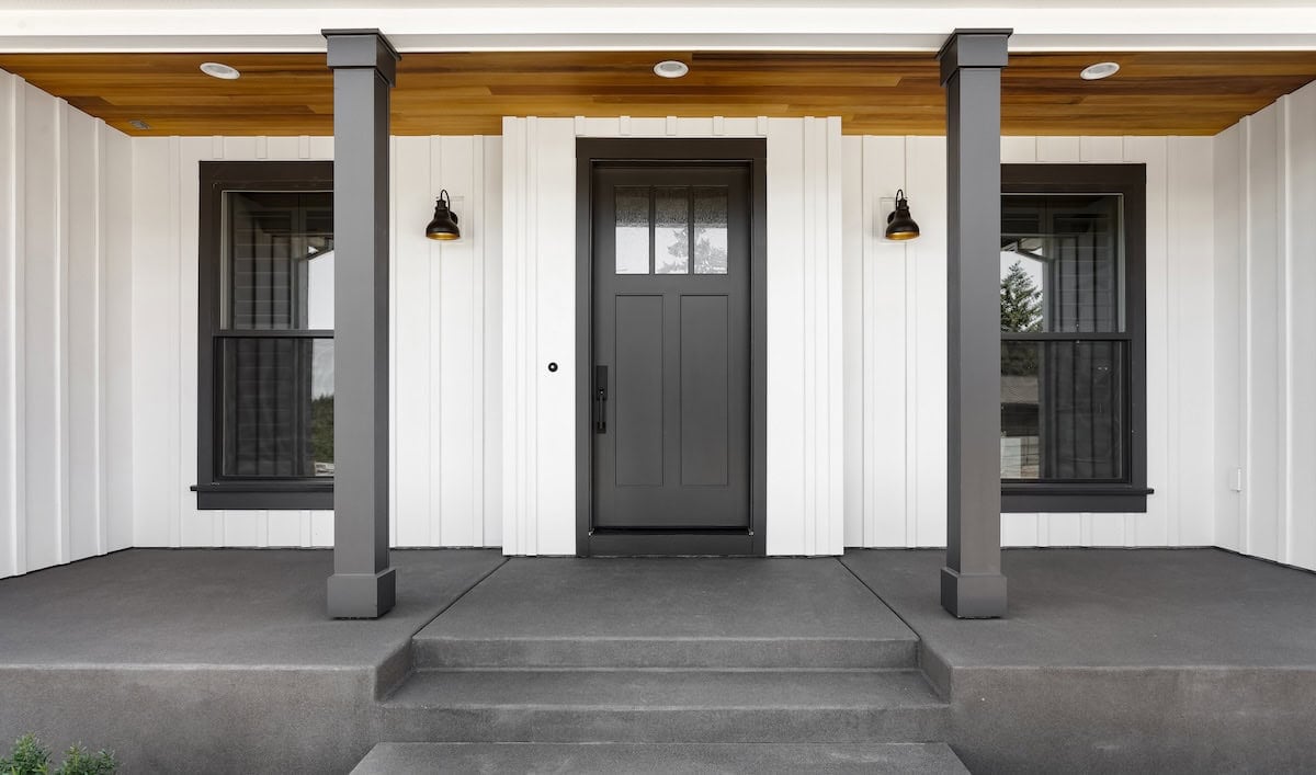 Front door to modern farmhouse home. Home exterior with white vertical wood siding and black front door.  Columns and sconce lights on both sides of door.