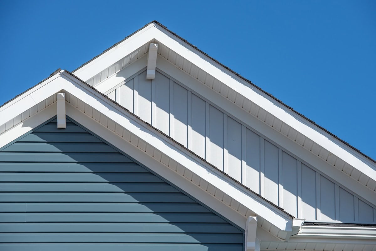 Double gable, with white decorative corbel, bracket, brace on a triangle gable roof, white soffit and fascia,  gray vertical and blue horizontal vinyl lap siding with blue sky background