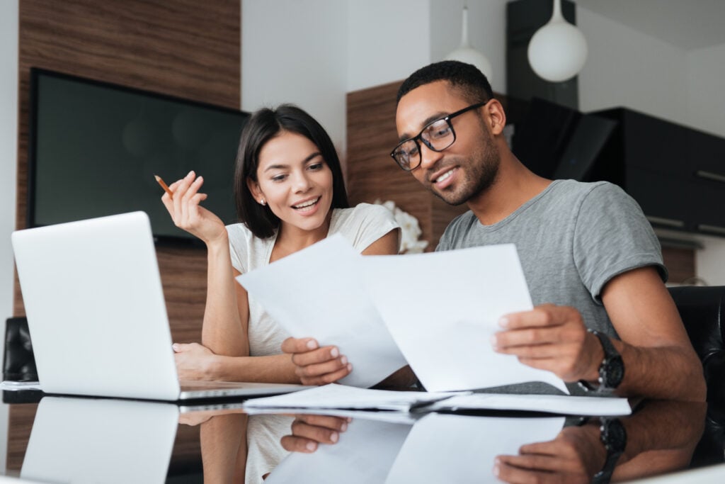 Photo of cheerful loving young couple using laptop and analyzing their finances with documents. Look at papers. roof financing