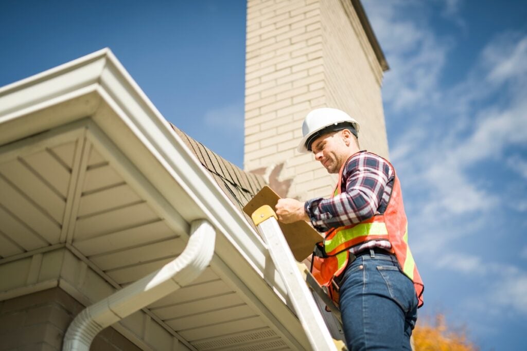 A man with hard hat standing on steps inspecting house roof