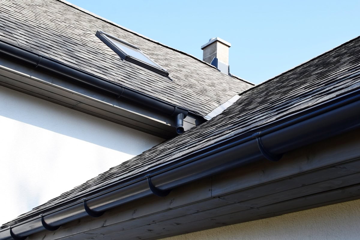 how to clean roof shingles Grey asphalt shingles roof construction with ridge ventilation on rooftop, wooden eaves with plastic rain gutter, window and chimney, copy space on white wall or blue sky.