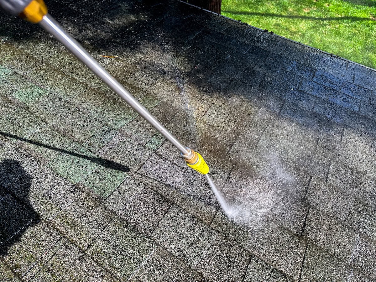 how to clean roof shingles A pressure washer is being used to clean the roof of a house on a clear day. Water sprays over the shingles, effectively removing dirt and debris from the surface